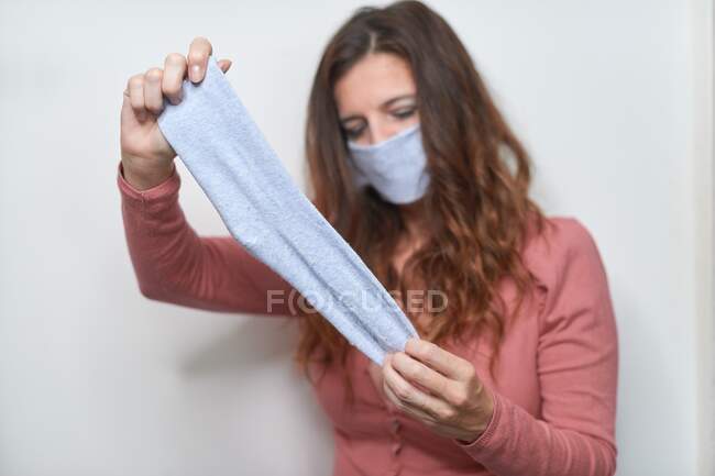 Mujer adulta con cabello castaño usando máscara respiratoria hecha a mano hecha de calcetín azul durante el período de cuarentena de la pandemia de coronavirus - foto de stock