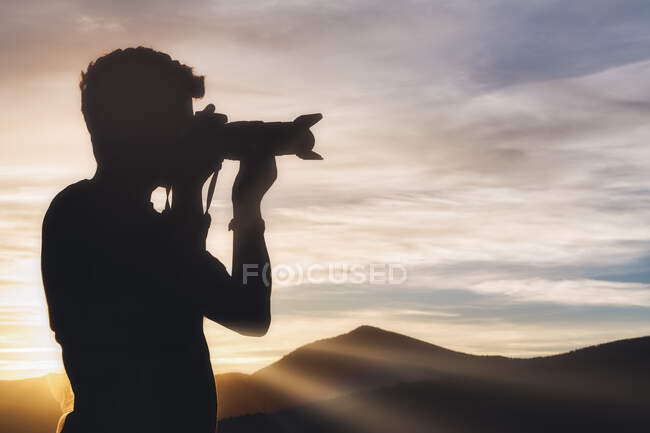 Vista lateral del joven viajero masculino en silueta de pie en la colina y tomando fotos de paisajes espectaculares de la cordillera al atardecer - foto de stock