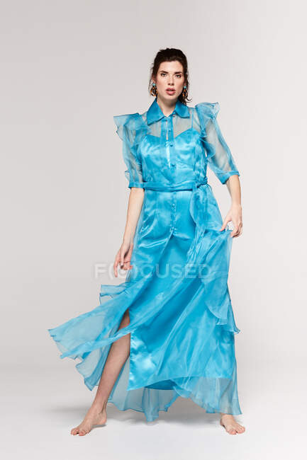 Full body of fashionable female with black hair in ponytail wearing light blue dress standing barefoot in white studio looking down — Stock Photo