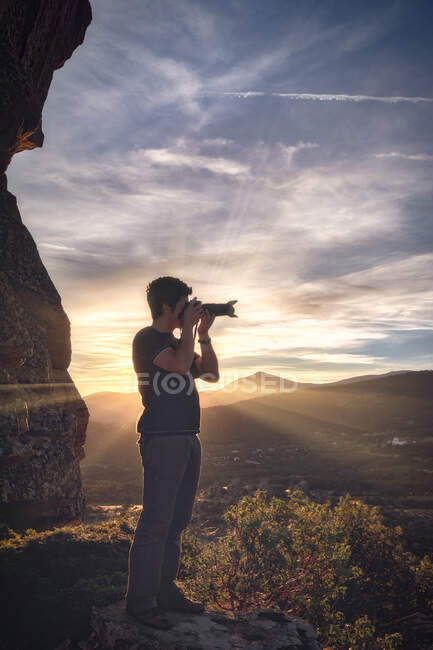 Vista lateral do jovem viajante masculino em roupas casuais em pé na colina e tirar fotos de paisagens espetaculares da cordilheira no pôr do sol — Fotografia de Stock