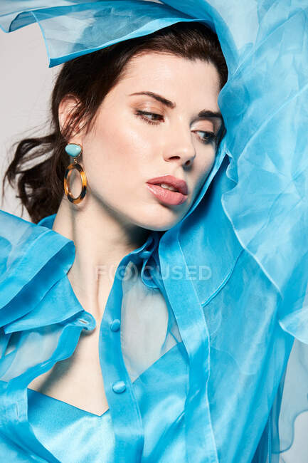 Alluring model wearing transparent blue blouse and covering face with textile while looking at camera in studio — Stock Photo