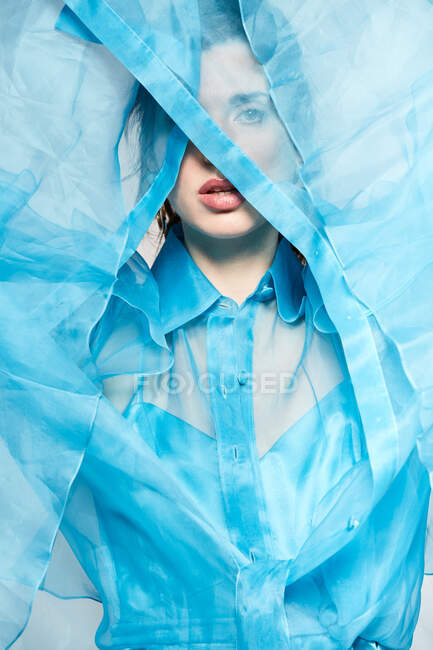 Приваблива модель в прозоро-блакитній блузці та покритті обличчя текстилем, дивлячись на камеру в студії — стокове фото