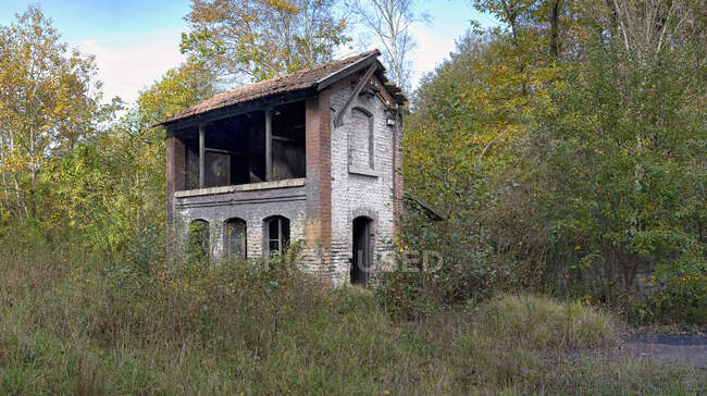 Shabby arruinado abandonado casa de ladrillo de dos pisos sin puerta y techo roto que se encuentra entre los árboles verdes en el día nublado en el campo - foto de stock