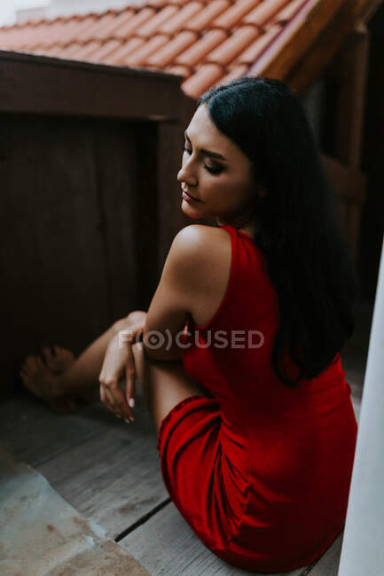 Alto ángulo de mujer descalza joven en vestido rojo de moda sentado piso de madera mientras descansa en la terraza - foto de stock