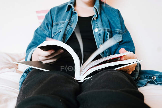Crop curious woman in black clothes and denim jacket flipping book with interest preparing to read while chilling alone on soft bed at home — Stock Photo