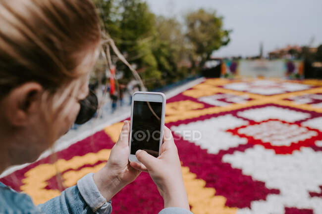 Crop faceless female traveler in jeans shirt and sunglasses taking photo of colorful big flowerbed on mobile phone while standing near fencing and looking at screen — Stock Photo