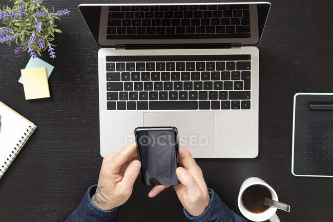 Зверху чоловік врожаю використовує смартфон, сидячи за столом з ноутбуком і чашкою кави вдома — стокове фото
