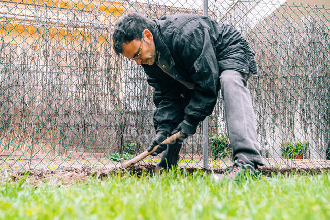 Вид сбоку на взрослого садовника мужского пола в рабочей одежде и перчатках, работающего с грабли во время обработки почвы перед посадкой в саду — стоковое фото