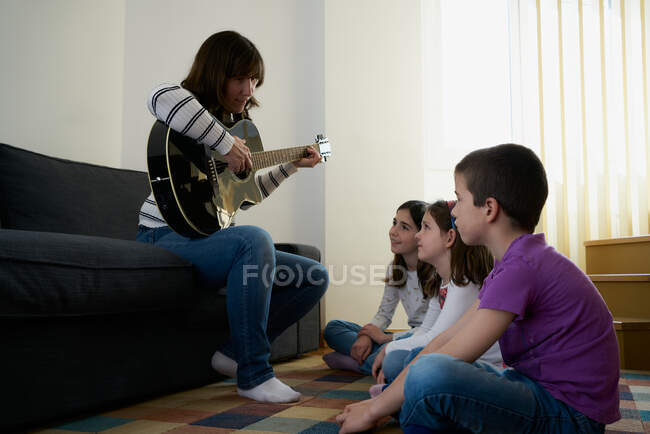 Mujer alegre tocando la guitarra para los niños en la sala de estar en casa - foto de stock