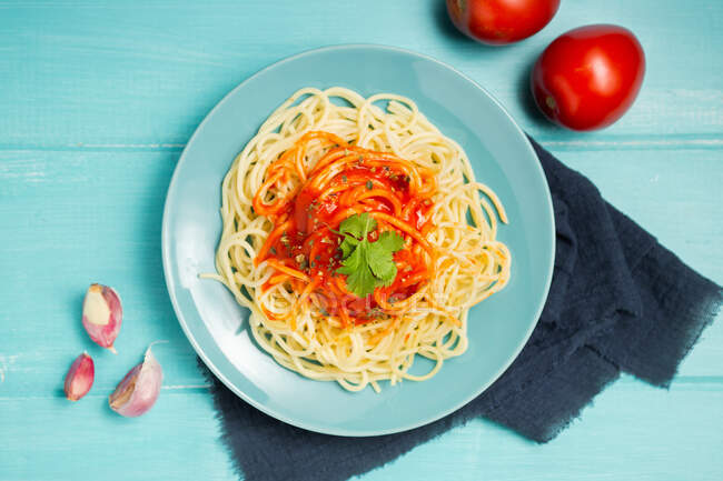 Vue de dessus de assiette en céramique bleue avec pâtes et sauce tomate décorée de persil et basilic servi entre gousses d'ail et couple de tomates sur fond bleu clair — Photo de stock