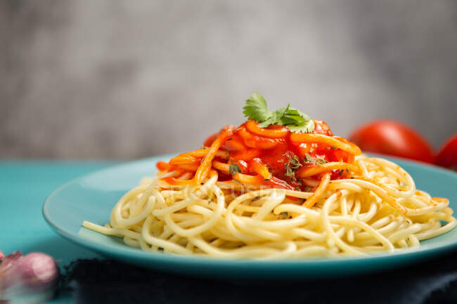 Голубая керамическая тарелка с макаронами и томатным соусом, украшенная петрушкой и базиликом, подается между зубчиком чеснока и парой помидоров на светло-голубом фоне — стоковое фото