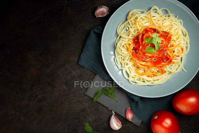 Vista superior de plato de cerámica azul con pasta y salsa de tomate decorado con perejil y albahaca servido entre dientes de ajo y un par de tomates sobre fondo oscuro - foto de stock