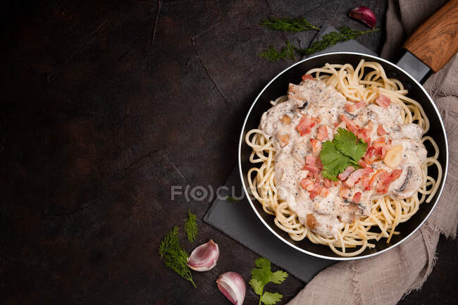 Composition de savoureux spaghettis aux tranches de jambon et champignons en sauce crémeuse cuite dans une casserole et placée sur une planche à découper en bois à table noire avec de l'ail et du lin gris de côté — Photo de stock