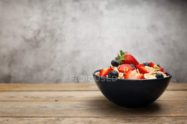 Von oben köstliche Frühstücksschüssel mit Cornflakes mit Erdbeeren und Blaubeeren auf Schneidebrett gelegt und mit Leinentuch und Beeren um Teller auf Holztisch mit grauem Hintergrund dekoriert — Stockfoto