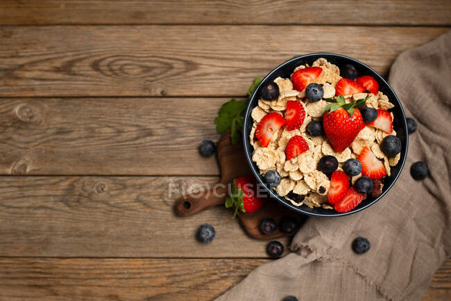 Vista superior de delicioso tazón de desayuno de hojuelas de maíz con fresas y arándanos colocados en la tabla de cortar y decorados con tela de lino y bayas alrededor del plato sobre fondo de madera - foto de stock