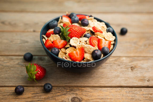 Blick von oben auf köstliche Frühstücksschüssel mit Cornflakes mit Erdbeeren und Blaubeeren auf Schneidebrett gelegt und mit Leinentuch und Beeren um Schale auf Holzgrund dekoriert — Stockfoto