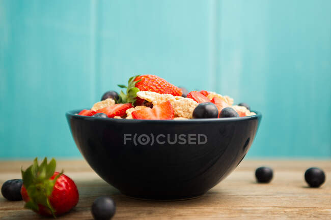 From above delicious breakfast bowl of corn flakes with strawberries and blueberries placed on cutting board and decorated with linen cloth and berries around dish on wooden table with blue background — Stock Photo