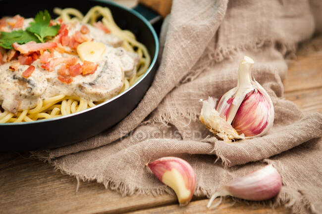 Spaghetti con fette di prosciutto e funghi in salsa cremosa cotti in padella e posti su tagliere di legno su un tavolo di legno con aglio e tessuto di lino da parte — Foto stock