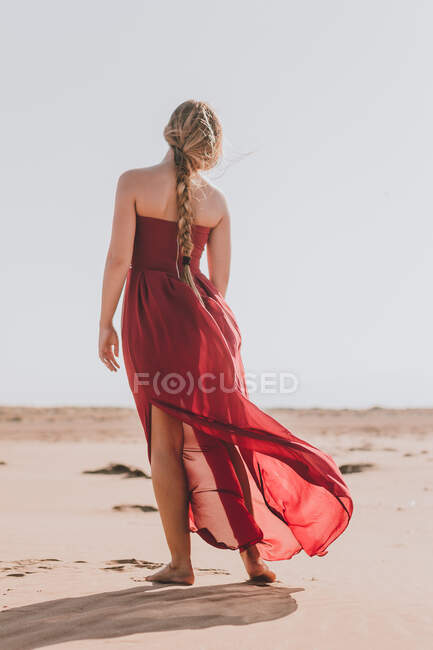 Vue arrière de jeune femme anonyme aux longs cheveux blonds portant une élégante robe rouge marchant sur le sable — Photo de stock