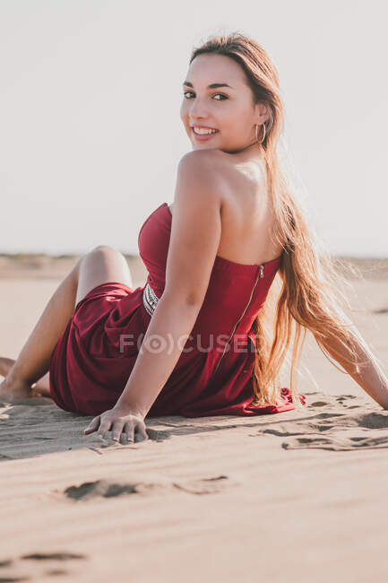 Jolie jeune femme aux longs cheveux blonds portant une robe rouge élégante assise sur la côte tout en regardant la caméra — Photo de stock