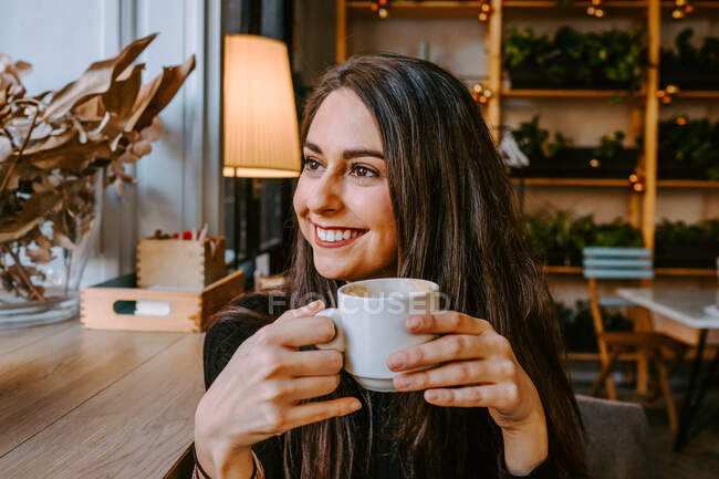 Optimistic young female smiling and looking away while enjoying fresh coffee in cozy cafeteria — Stock Photo