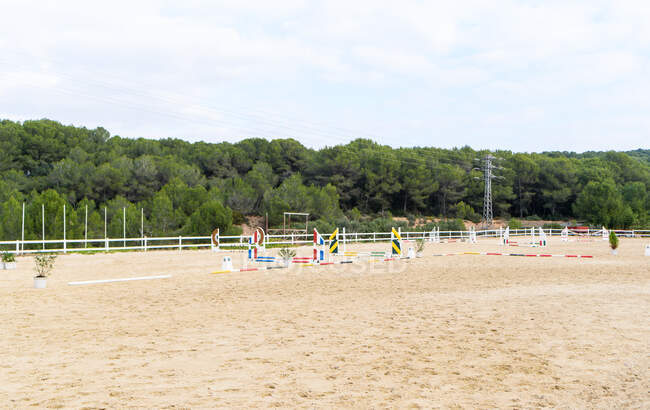 Multicolores obstáculos instalados en el suelo arenoso en el paddock de la escuela de equitación cerca de árboles verdes en el día nublado en el campo - foto de stock