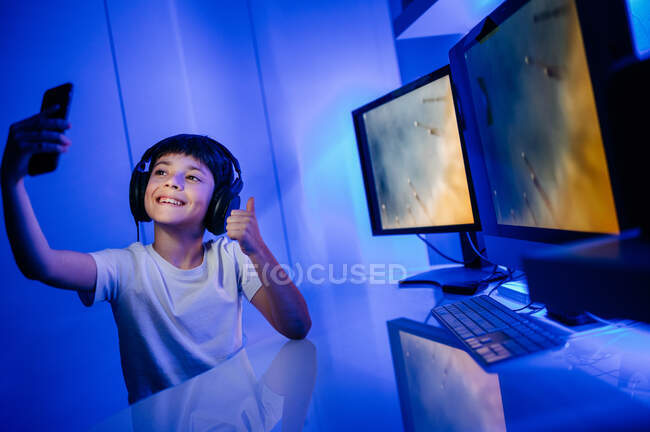 Content male child in headphones taking selfie on cellphone while sitting with thumb up at table with desktop computer in room — Stock Photo