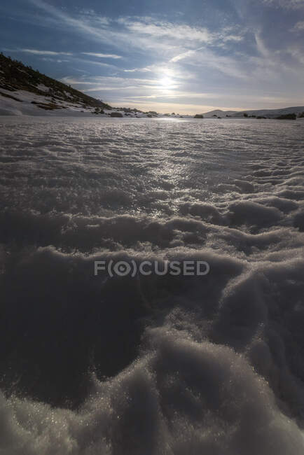 Majestuosa vista del lago helado nevado ubicado en medio de la zona montañosa nevada contra el cielo soleado nublado en el frío día de invierno - foto de stock