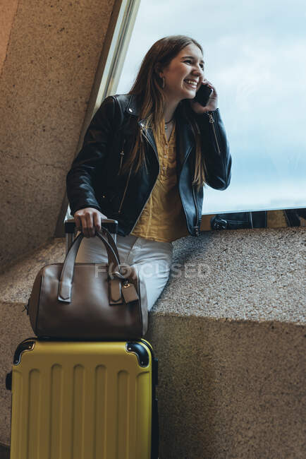 Crop female traveler in casual clothes with suitcase and bag standing by window leaning on wall and talking on smartphone at airport — Stock Photo