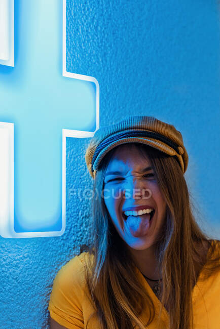 Mujer joven feliz en camiseta amarilla y gorra de moda haciendo mueca divertida y mostrando la lengua contra la pared azul con signo de neón de cruz médica - foto de stock