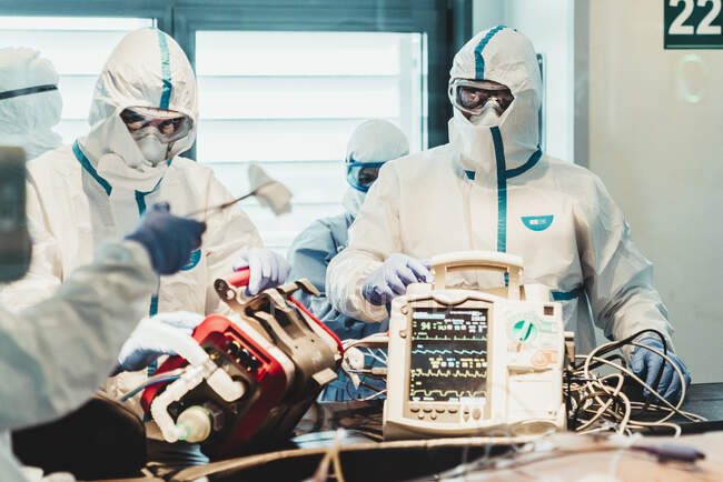Grupo de médicos profesionales con máscaras protectoras y trajes de pie cerca de la mesa de operaciones con equipo y preparándose para la operación en la clínica moderna - foto de stock
