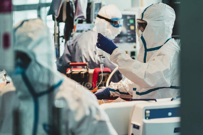 Vista lateral del médico que usa uniforme protector y guantes de látex comprobando el sistema de infusión mientras trabaja en una clínica moderna - foto de stock
