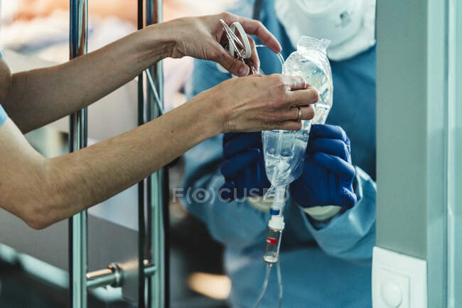 Anonymous crop medical nurse giving infusion system to doctor in protective costume and mask in modern clinic during work — Stock Photo
