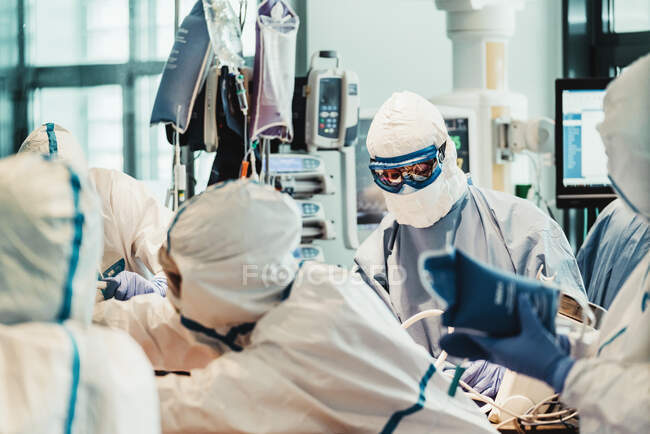Grupo de médicos profesionales con máscaras protectoras y trajes de pie cerca de la mesa de operaciones con equipo y preparándose para la operación en la clínica moderna - foto de stock