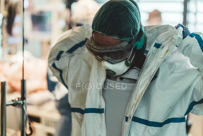 Cirujano cansado quitándose la máscara protectora y el uniforme mientras sale del quirófano después de una operación difícil en la clínica moderna - foto de stock