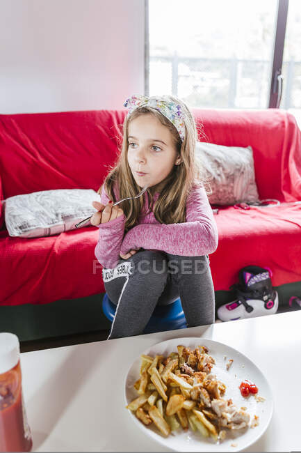 Девушка в повседневной одежде ест вкусное блюдо и смотрит в сторону, сидя рядом с диваном и обедая дома — стоковое фото