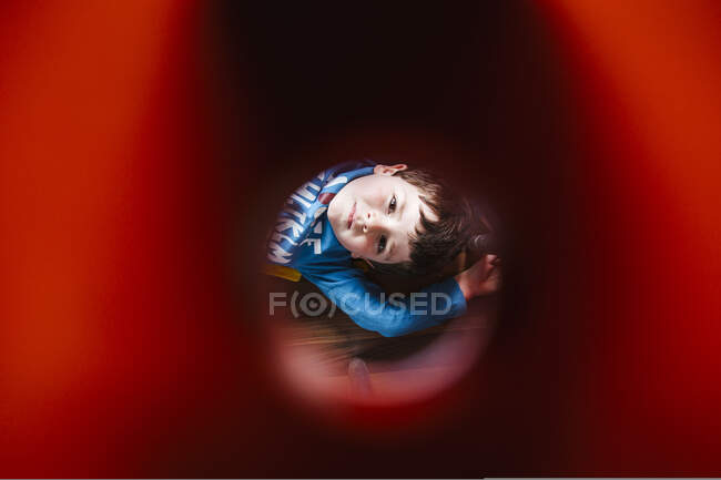 Маленький хлопчик лежить на підлозі і дивиться на камеру через червону трубку, граючи вдома — стокове фото