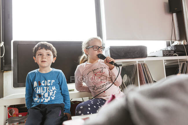 Дівчина в окулярах співає караоке пісню, сидячи біля брата в затишній кімнаті у вихідний день вдома — стокове фото