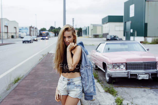 Sinnliches blondes Mädchen, das auf dem Bürgersteig steht und in die Kamera schaut, in der Nähe eines klassischen rosa Autos — Stockfoto