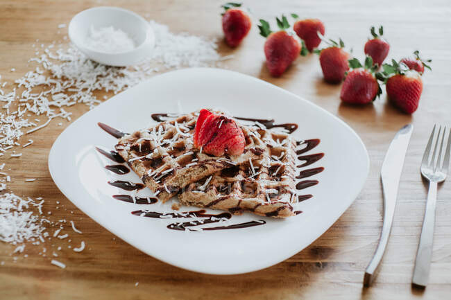 Gaufres douces et fraises servies dans une assiette avec sirop de chocolat délicieux et flocons de noix de coco sur une table en bois près de l'argenterie — Photo de stock