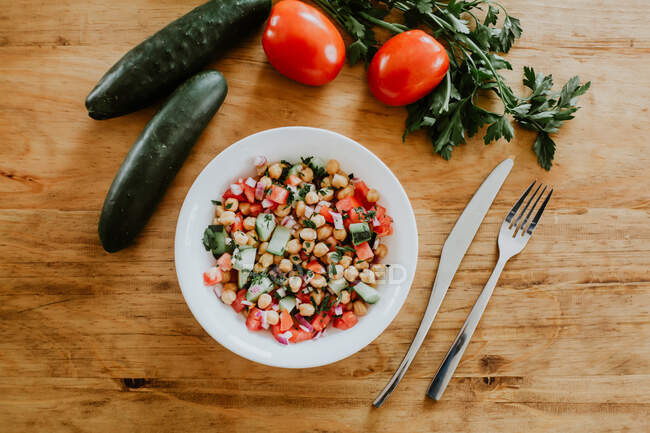De arriba cuenco de pepino sano y ensalada de tomate con garbanzos y perejil colocado en la mesa de madera cerca de tenedor y cuchillo - foto de stock