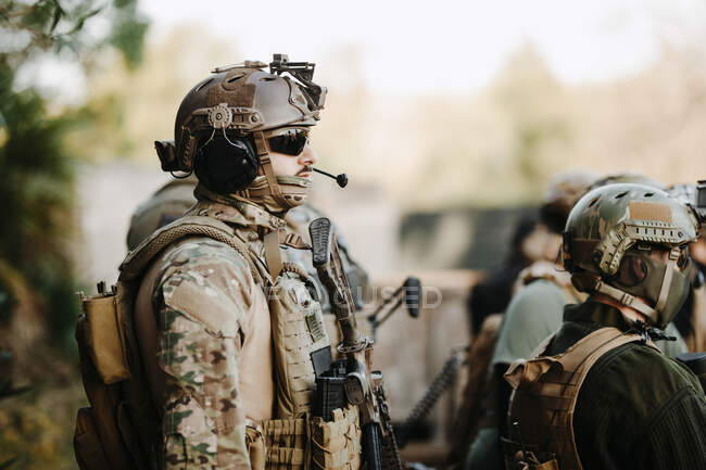 Vista lateral del hombre adulto en uniforme militar y con pistola airsoft escuchando briefing con escuadrón - foto de stock