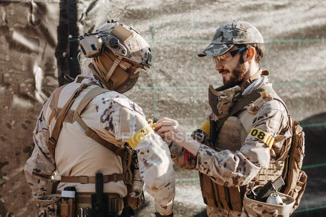Bearded man in camouflage fastening identification band of arm of squadmate while preparing to play airsoft game together — Stock Photo
