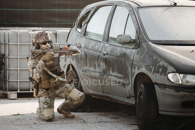 Homem adulto em uniforme militar apontando e disparando arma enquanto deitado no chão perto do veículo durante a partida airsoft — Fotografia de Stock