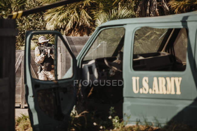 Homem em camuflagem escondido atrás de barreira de madeira e disparando airsoft arma atrás de veículo militar enquanto joga jogo tático — Fotografia de Stock
