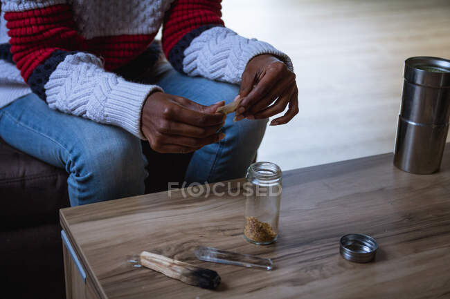 Женщина в повседневной одежде сидит на диване за столом с банкой сухого табака и катает сигарету дома — стоковое фото