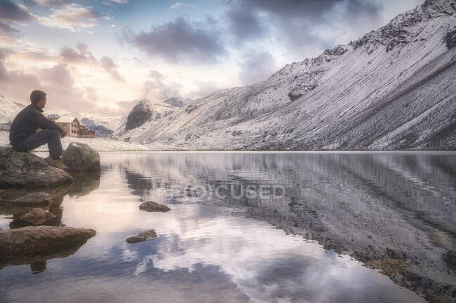Vue latérale du mâle adulte assis sur la pierre près d'un lac calme et d'une montagne enneigée et admirant le ciel nuageux au coucher du soleil lors de la visite du parc national suisse en Suisse — Photo de stock