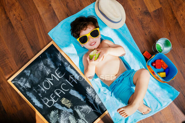Desde arriba vista de niño alegre en gafas de sol amarillas y trajes de baño azules comiendo manzana mientras está acostado en la toalla en el suelo tener casa playa en cuarentena - foto de stock