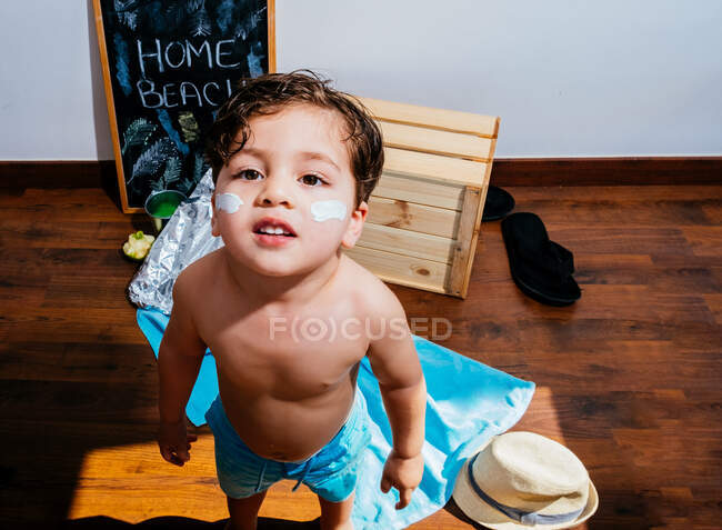 Зверху позитивний маленький хлопчик у блакитному купальнику стоїть на дерев'яній підлозі, де пляж вдома знаходиться на карантині і дивиться на камеру — стокове фото