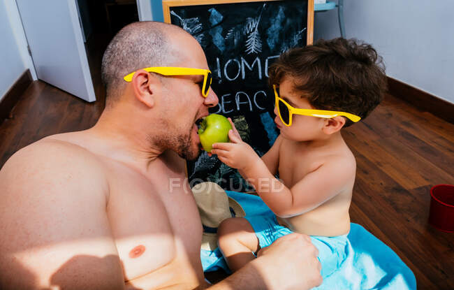 Сверху вид веселого мальчика в желтых солнцезащитных очках и синих купальниках, улыбающегося во время кормления отца яблоком, лежащим на полотенцах вместе, имеющим пляж дома — стоковое фото